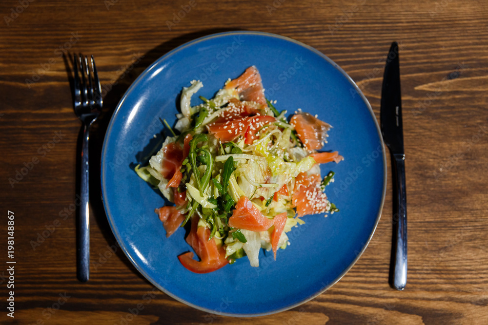 A horizontal image of salad with salmon, lettuce, arugula, tomatoes, sesame seeds. Served on a bright blue plate. A horizontal image of salad with salmon, lettuce, arugula, tomatoes, sesame seeds. Served on a bright blue plate.