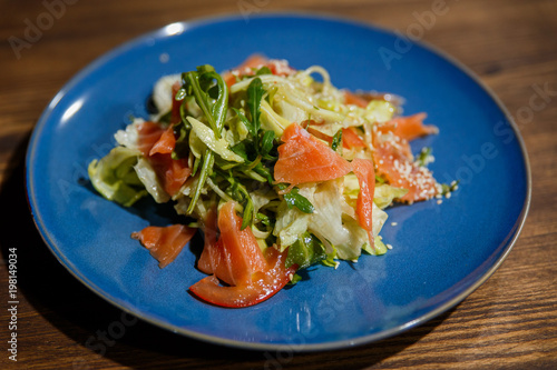 A horizontal image of salad with salmon, lettuce, arugula, tomatoes, sesame seeds. Served on a bright blue plate.