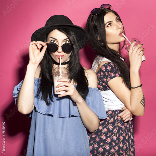 Two caucasian brunette hipster woman in casual stylish outfit having fun drinking milkshakes with straws. They standing on a bright pink background. Cheerful, happy emotions