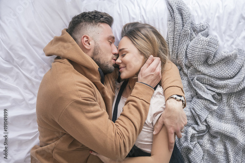 Stylish hipster couple of man and woman sitting on bed in their home in cozy casual clothes. They hug and love each other and smile happily, resting together. Space for text