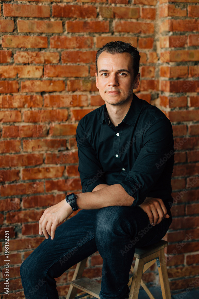 Cute young man in black shirt sitting on chair in studio near brick wall and looking at camera.