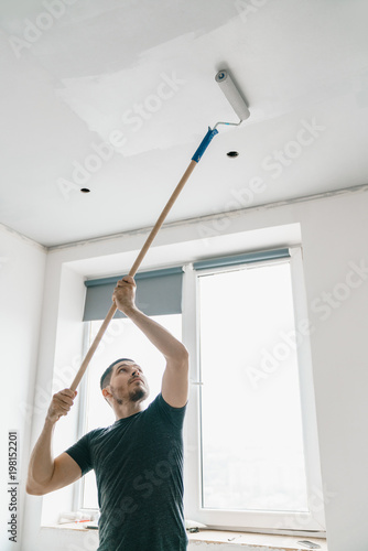 a man with a roller in his hand on a long stick paints the ceiling in gray at the window. Repair in a bright room.