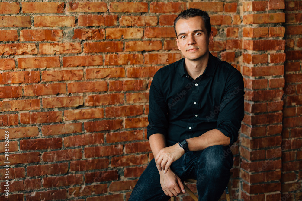 Cute young man in black shirt sitting on chair in studio near brick wall and looking at camera. Cute young man in black shirt sitting on chair in studio near brick wall and looking at camera.