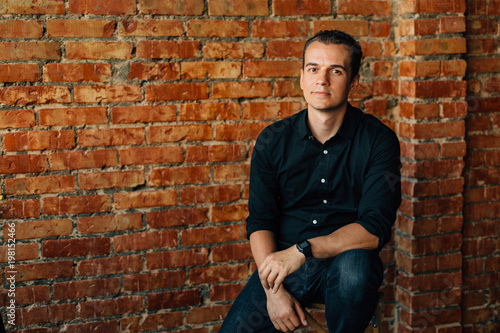 Cute young man in black shirt sitting on chair in studio near brick wall and looking at camera.