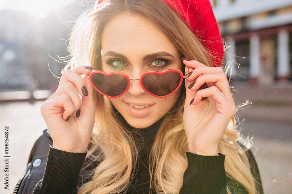 Naklejka premium Close-up portrait of attractive blond female model dressed red hat and stylish glasses in sunlight. Photo of happy caucasian girl in sunglasses posing outside.