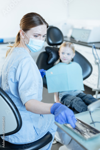 Thorough checkup. Pleasant young female dentist carrying out a preliminary checkup of her little patient and choosing a necessary instrument for that purpose