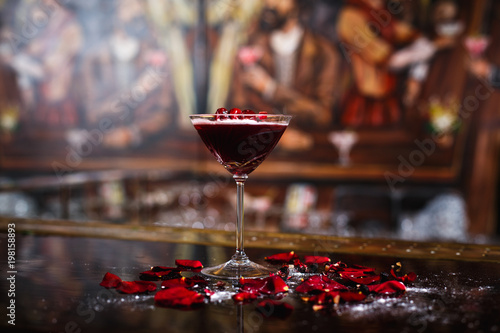 A horizontal photo of a cocktail glass with a deep red alcoholic drink, garnished with cherry, rose petals and powdered sugar. Selective focus. photo