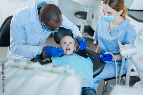 Coordinated work. Pleasant male dentist carrying out an examination of her little patients oral cavity while his nurse holding a saliva ejector