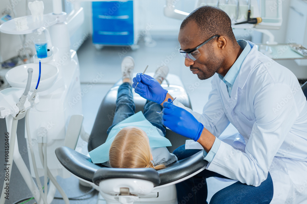 Fototapeta premium Best teeth care. Pleasant caring male dentist using a dental probe and a mouth mirror and checking the teeth of a little girl lying in the chair