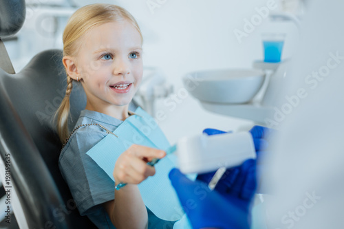 Diligent patient. Adorable little girl sitting in a dentist chair and learning how to brush teeth correctly, practicing on a dental model