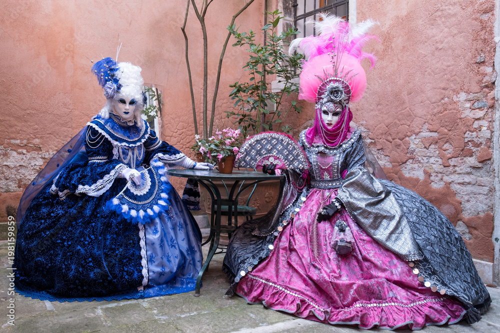 Two masked ladies in blue and pink costumes sitting at a table in a courtyard with an old terracotta wall behind them at Venice Carnival (Carnivale di Venezia) Two masked ladies in blue and pink costumes sitting at a table in a courtyard with an old terracotta wall behind them at Venice Carnival (Carnivale di Venezia)