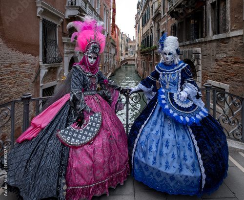Two women in pink and blue costumes with fans and ornate painted feathered masks at Venice Carnival. Women are standing on a bridge with canal in background.