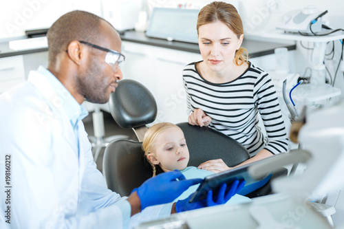 Have a look. Professional male dentist showing a dental X-ray on the tablet to his little patient and her mother while they looking at it with worried expression