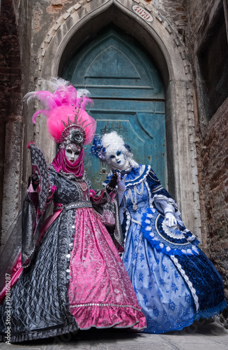Two women in masks and ornate blue and pink costumes standing in front of an old blue door at the end of an alley in Venice during the carnival (Carnivale di Venezia).