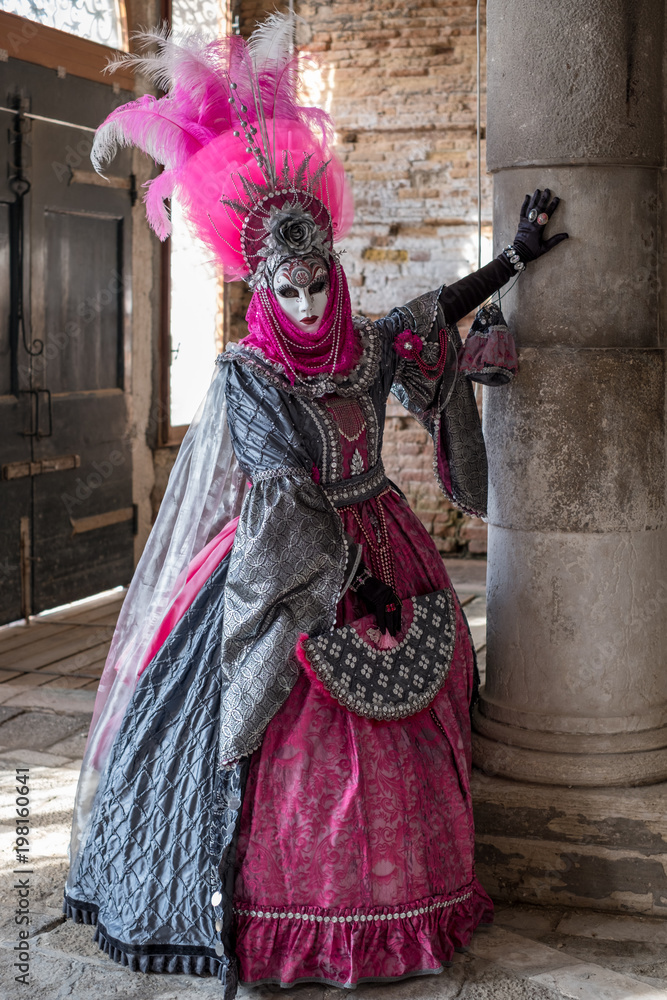  Masked lady in ornate pink costume dress and mask standing next to pillar at Venice Carnival. Lady has feathered plumes, holds a decorated fan and is wearing black gloves