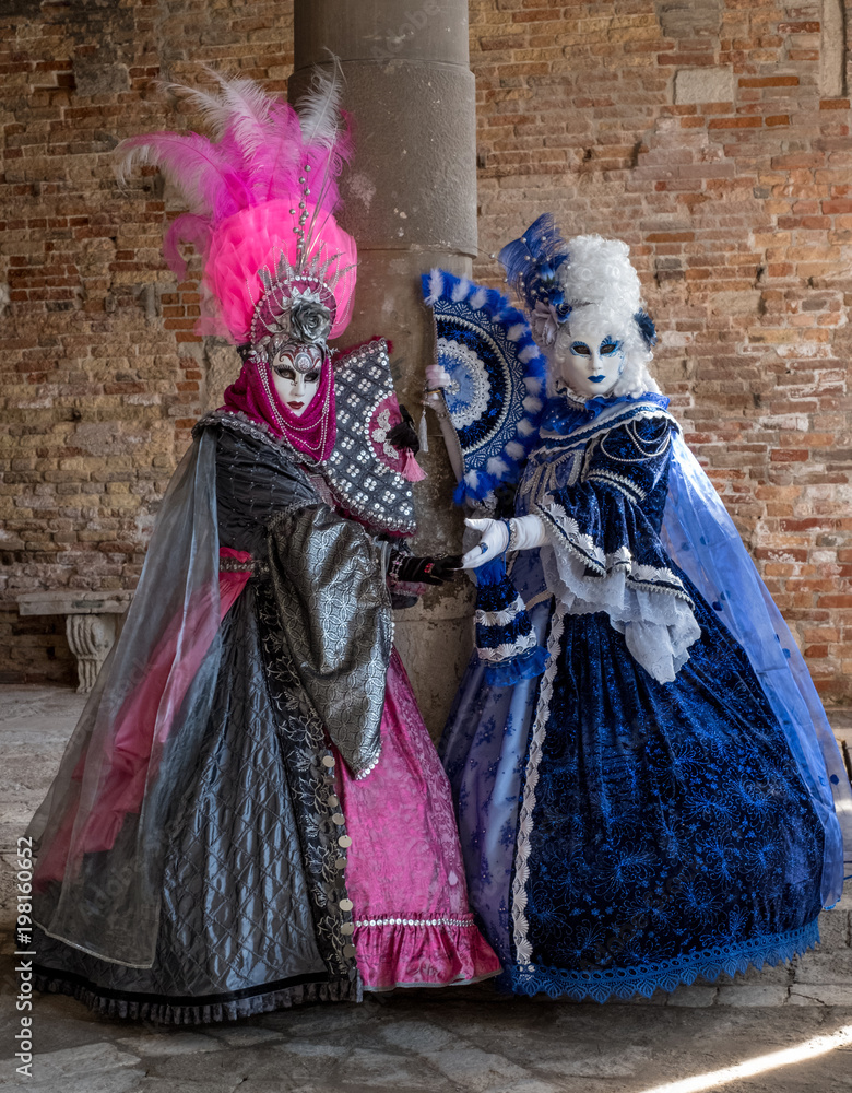 Two women in masks and ornate blue and pink costumes and decorative fans, standing against an old brick wall. 