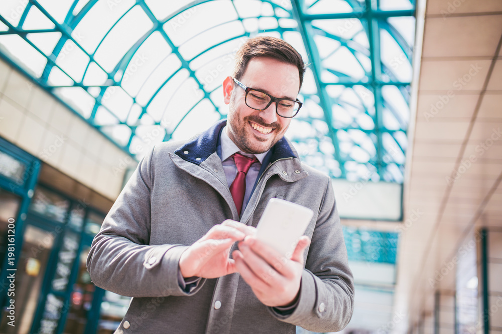 Fototapeta premium Businessman using mobile phone outside of office buildings in the background. Young caucasian man holding smartphone for business work.