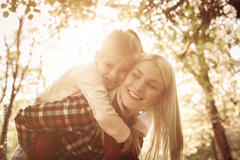 Fototapeta premium Smiling mother carrying her daughter on piggyback and enjoying in nature.