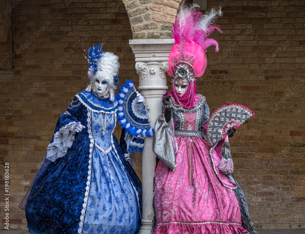Masked women in ornate blue and pink costumes standing next to stone column in a courtyard during the carnival (Carnival di Venezia)