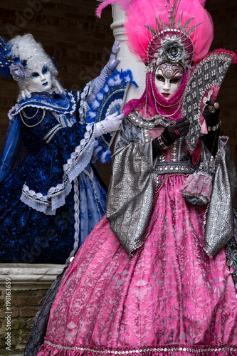 Masked women in ornate blue and pink costumes standing next to stone column in a courtyard during the carnival (Carnival di Venezia)