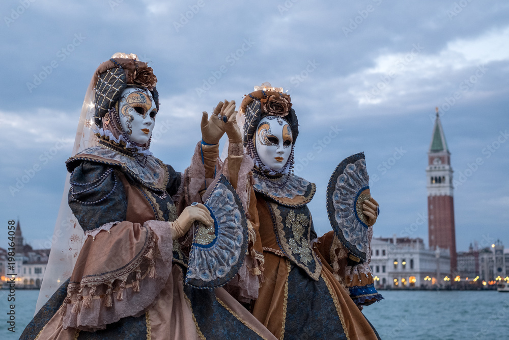 Obraz premium Venice Italy, February 2018. Two women in traditional costumes and masks, with decorated fans, standing in front of the Grand Canal with San Marco in the background, during the Venice Carnival
