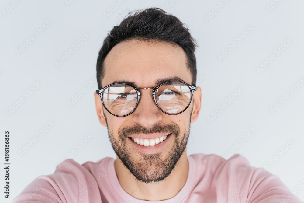 Closeup portrait of handsome young happy smiling bearded male model look at  the camera, wears round trendy glasses with toothy smile. Cheerful man in  spectacles, pink clothes advertises in studio wall Stock