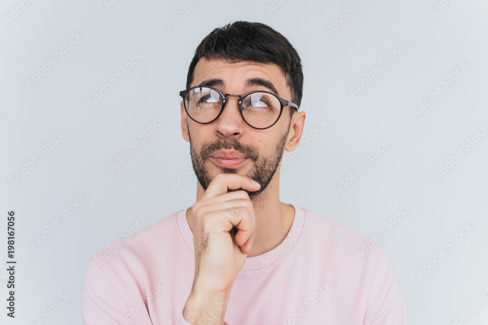 Horizontal portrait of handsome pensive male keeps hand under chin, looking up, isolated over white studio background. Successful bearded man thinking about plans, wears pink clothes. People concept