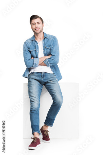 young smiling man in denim leaning on white cube, isolated on white photo