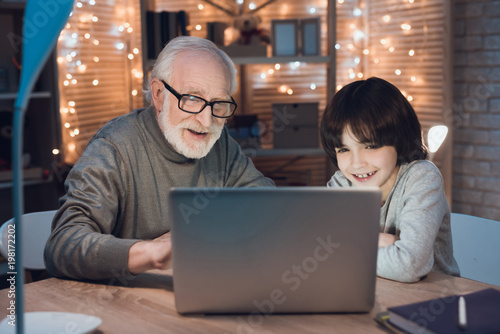 Grandfather and grandson are watching video on laptop at night at home.