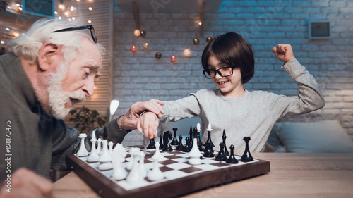 Grandfather and grandson are playing chess together at night at home. Boy is winning.