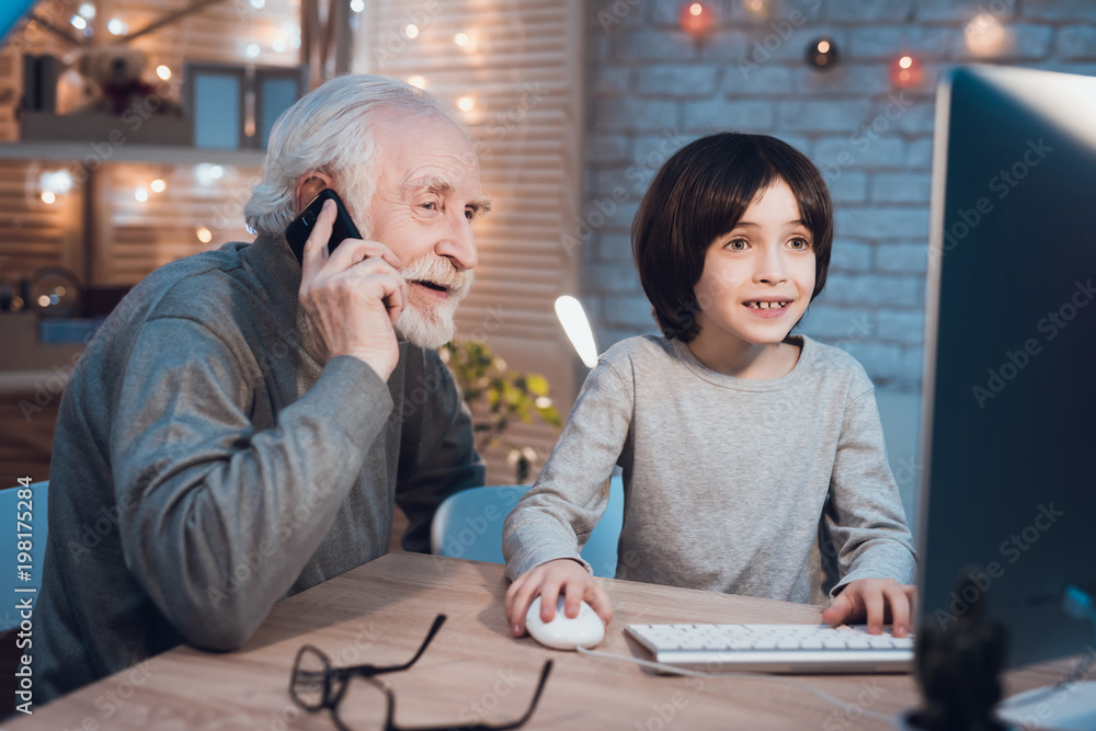 Grandfather and grandson are playing games on computer at night at home. Granddad is on phone. Grandfather and grandson are playing games on computer at night at home. Granddad is on phone.