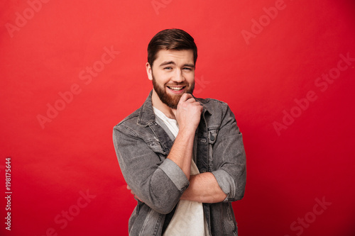 Portrait of affable kind man 30s in jeans jacket posing on camera touching chin with beautiful smile, isolated over red background © Drobot Dean