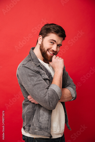 Portrait of pleased young man 30s in jeans jacket posing on camera touching chin with candid smile, isolated over red background