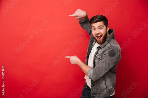 Photo of agitated man wih beard in casual clothing pointing fingers on copyspace text or product with surprise and smile, isolated over red background photo