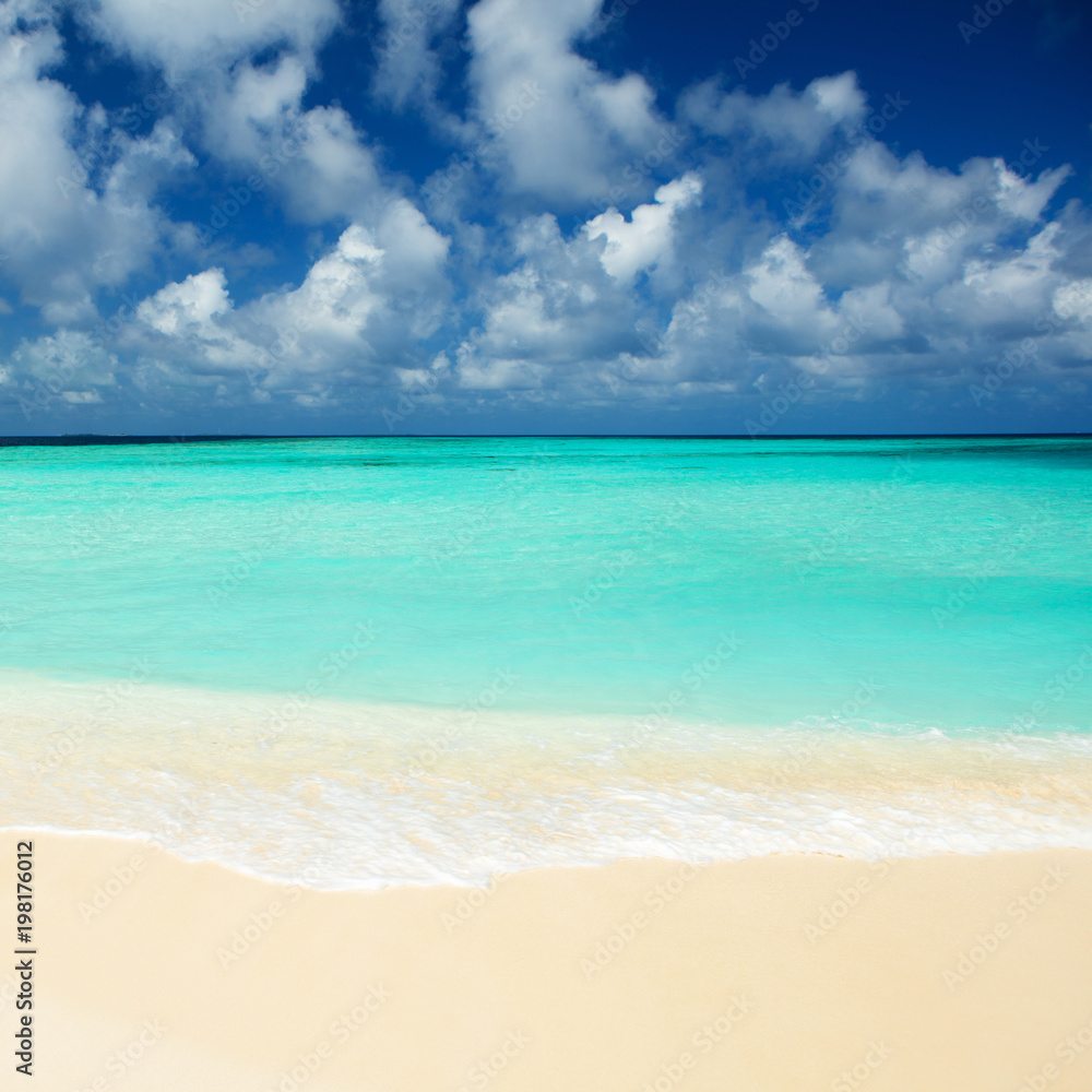 Tropical beach. Ocean waves and cloudy sky background. White sand and crystal-blue sea. Ocean water nature, beach relax. Summer sea vacation. Maldives islands sea background