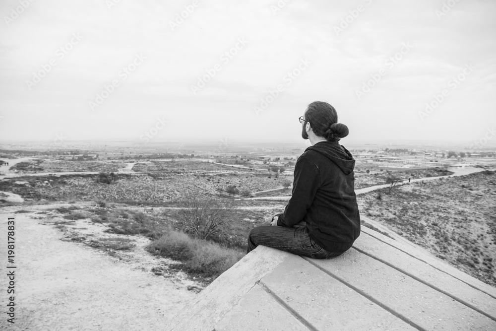 Side view unidentified young man in casual clothes and glasses sits on the high rock and looks at beautiful desert