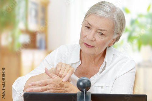 Canvastavla Senior woman in front of a computer with an attached camera showing her doctor a mole on her hand during video call, teledermatology concept