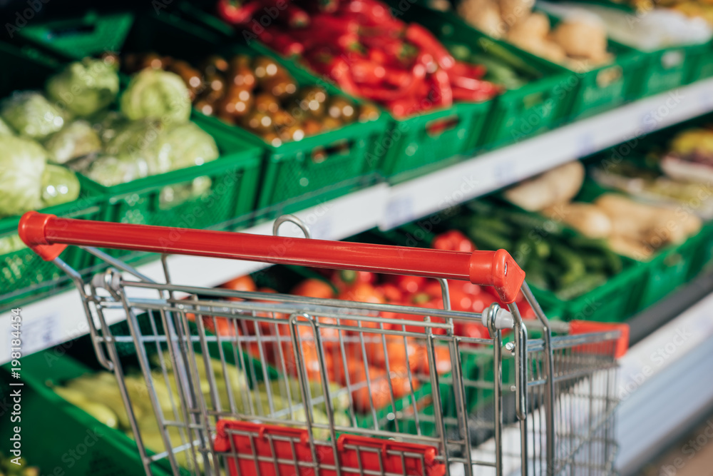 selective focus of empty shopping cart and fresh vegetables in supermarket