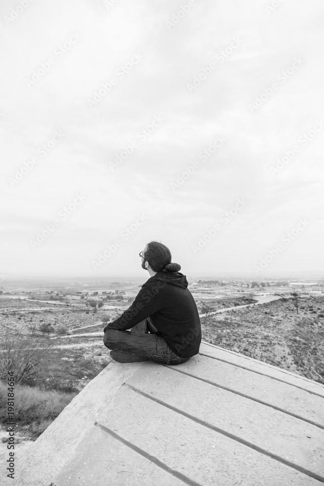 Side view unidentified young man in casual clothes and glasses sits on the high rock and looks at beautiful desert Side view unidentified young man in casual clothes and glasses sits on the high rock and looks at beautiful desert