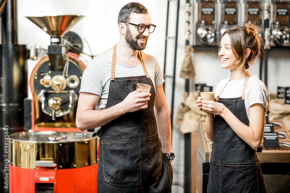 Portrait of a couple of baristas in uniform standing together in the coffee shop with coffee roasting machine on the background
