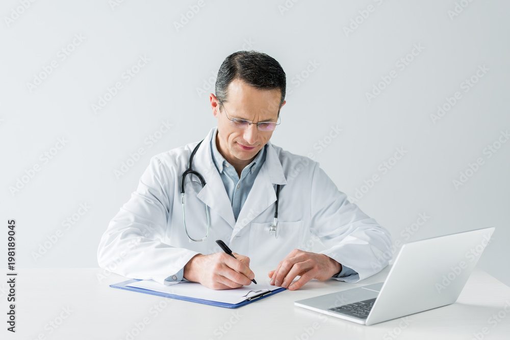serious adult doctor sitting at workplace and writing diagnosis in clipboard isolated on white serious adult doctor sitting at workplace and writing diagnosis in clipboard isolated on white