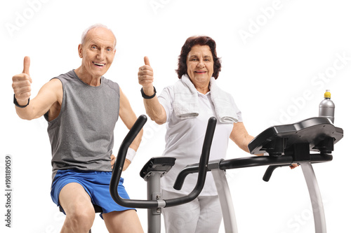 Elderly man on a stationary bike and an elderly woman on a treadmill holding their thumbs up