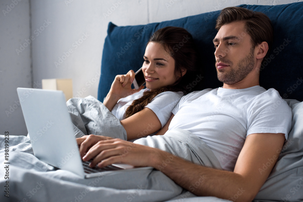 Fototapeta premium Young man and woman using digital devices while lying in bed