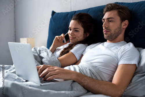 Schilderij op canvas Young man and woman using digital devices while lying in bed