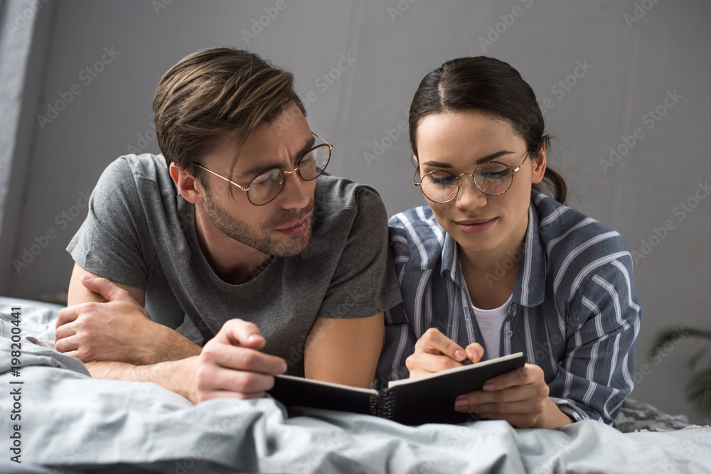 Thoughtful man looking at woman writing in notepad while lying in bed Thoughtful man looking at woman writing in notepad while lying in bed
