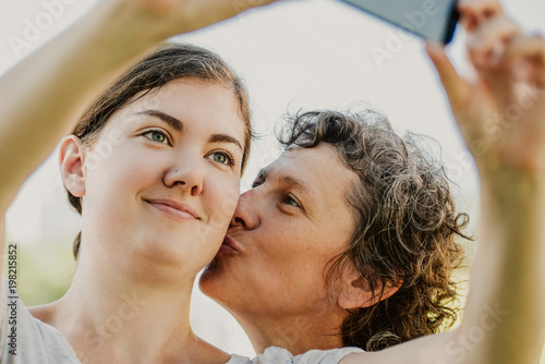 Portrait of smiling teenage Caucasian girl taking selfie while her senior mother kissing her