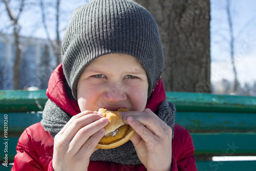 Small boy is eating burger outdoor.  Hungry child eats burger in the park sunny spring day. Concept of childhood.