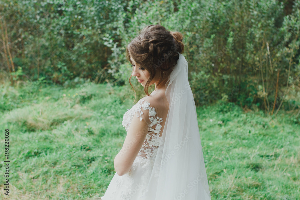 Beautiful bride with elegant simple hairdo is standing in the forest. Low open back white dress with flowers and tulle veil. Stylish hairstyle in outdoors romantic wedding photo. Bridal portrait.