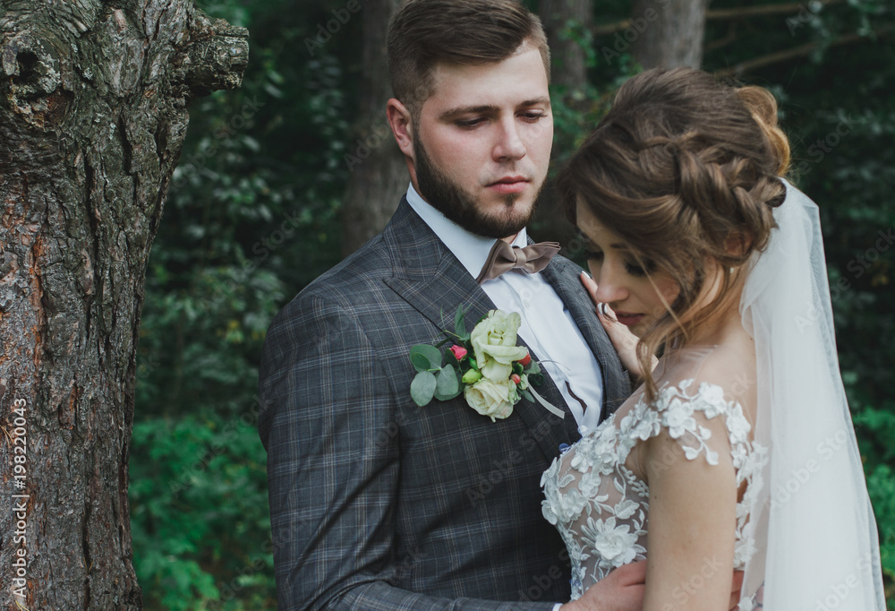 Beautiful wedding couple in the forest. The bride with tulle veil and open low back elegant dress is touching the groom in bow tie. Wedding buttonhole and checkered suit. Rustic outdoors photo. Beautiful wedding couple in the forest. The bride with tulle veil and open low back elegant dress is touching the groom in bow tie. Wedding buttonhole and checkered suit. Rustic outdoors photo.