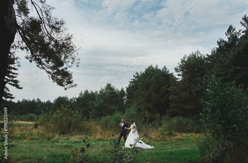 Beautiful wedding couple in the forest. The bride with tulle veil and trained open low back dress is walking with her groom in checkered suit. Rustic outdoors love story photo.
